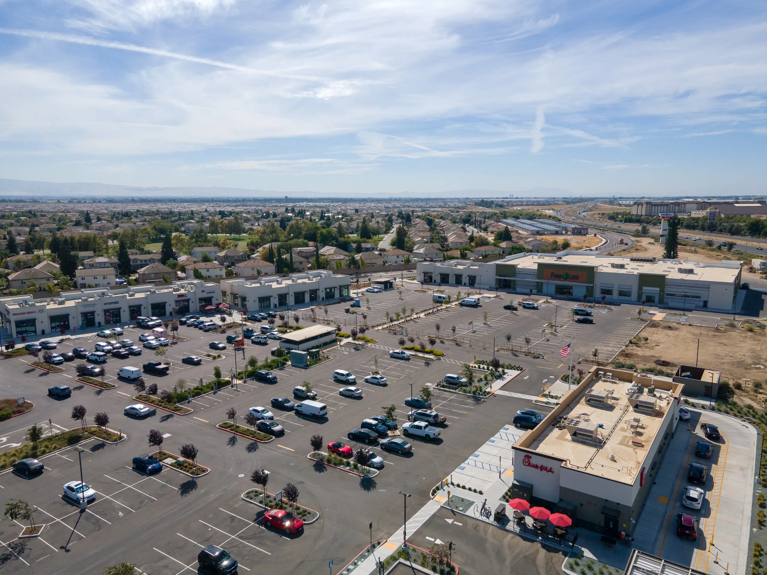 Aerial view across the main parking field with Chick-fil-A in the foreground, Shops A to the left, and Food 4 Less and Shops H beyond